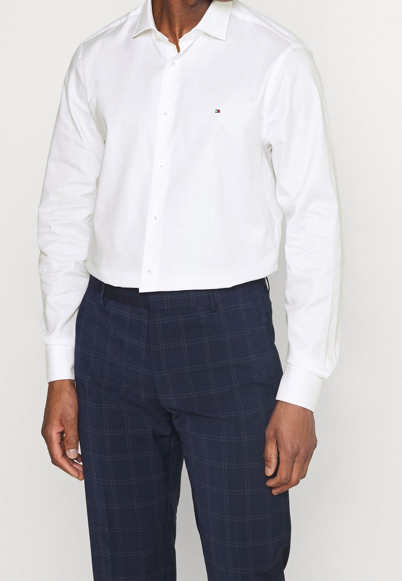 Camicetta bianca con colletto a punta e fronte con bottoni, indossata con pantaloni a quadri blu navy. Presenta un piccolo logo sul petto.