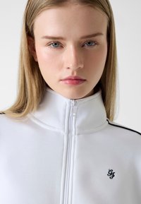 Weiße Jacke mit Reißverschluss und hohem Kragen, mit schwarzen seitlichen Akzenten und besticktem Logo. Glatte Textur, sauberes Design, Frontalansicht.