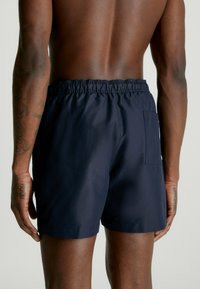 Costumi da bagno blu navy con cintura elastica e una tasca sul retro. Il tessuto sembra liscio e leggero.