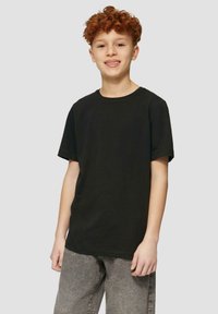 Junge mit lockigem roten Haar, der ein schwarzes T-Shirt mit kurzen Ärmeln und graue Jeans trägt, lächelt und steht vor einem schlichten hellgrauen Hintergrund.