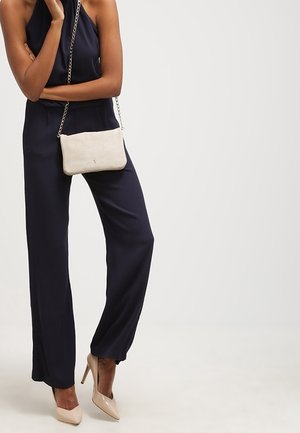 Combinaison bleu marine avec encolure halter, en tissu doux, coupe ample. Sac bandoulière beige accentué de chaînes. Talons hauts de couleur claire.