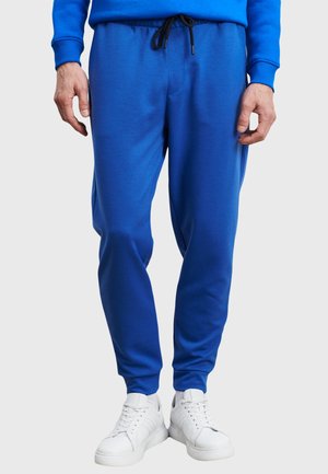 Mann in leuchtend blauen Jogginghosen mit schwarzen Kordeln und weißen Sneakers, der vor einem schlichten hellen Hintergrund steht.