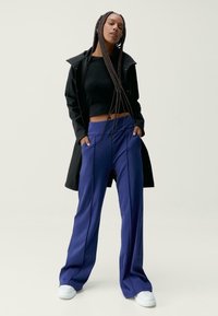 Črn krojen pulover, dolge temno modre hlače in črn plašč. Ima ohlapen kroj, mehak materiali in bele superge.