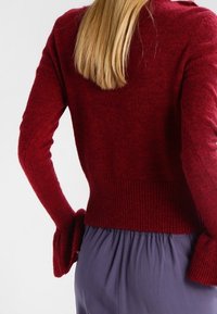 Roter, strukturierter Pullover mit geripptem Saum und weit ausgestellten Ärmelbündchen. Weiches Material, mittellanges Design, von hinten gezeigt mit grauen Hosen.