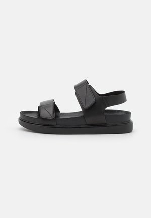 Sort åben sandal med to justerbare remme og en flad sål på hvid baggrund.