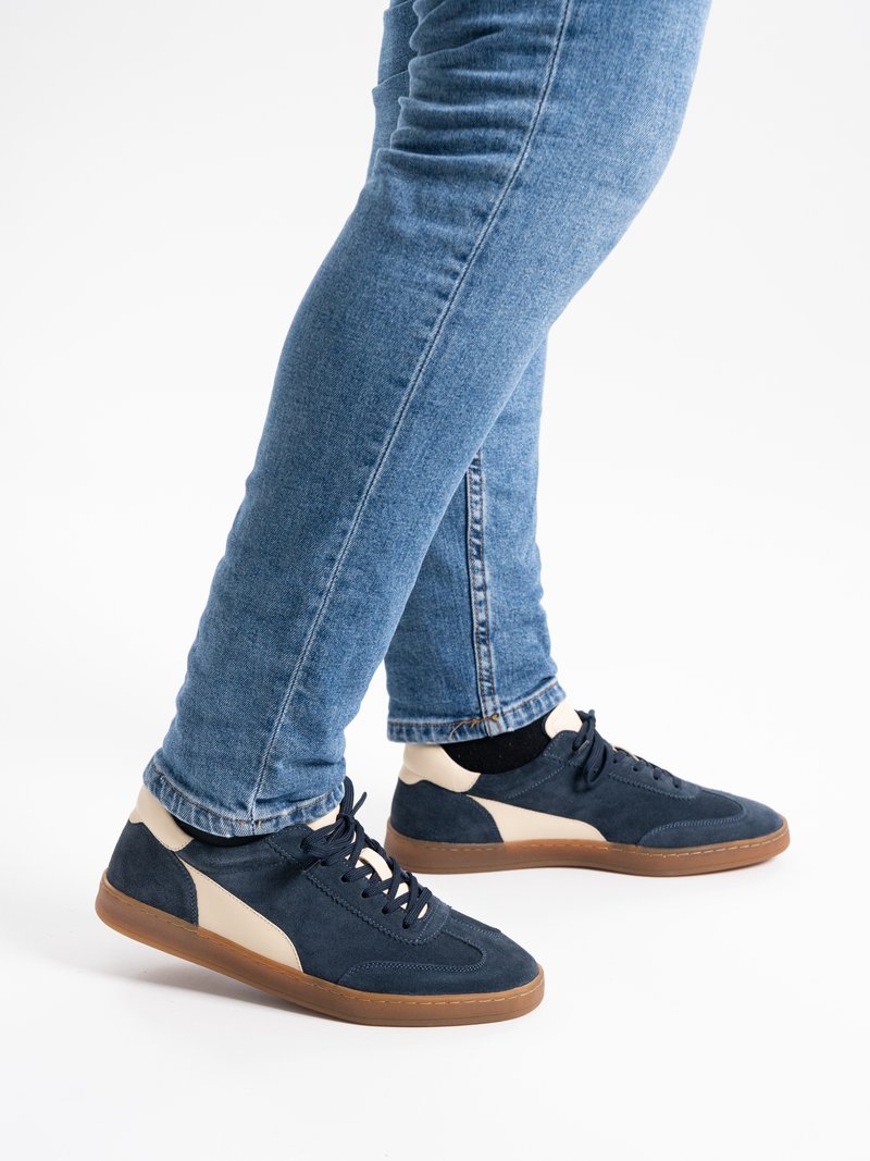 Legs wearing blue jeans and navy blue and beige sneakers with brown soles standing on a white background.