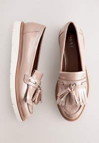 Mocassins en cuir verni beige clair, ornés d'une frange décorative et de détails à pampilles, avec une semelle épaisse blanche et des surpiqûres accentuées marron.
