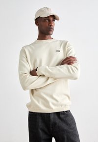 Sudadera de manga larga color crema con puños y cuello de canalé, que presenta un pequeño logo negro "BOSS" en el pecho; llevada con pantalones oscuros.