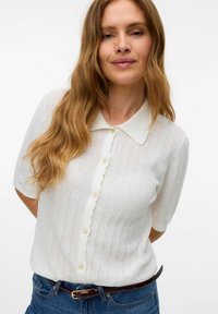 Blusa corta a maniche corte bianca in maglia con colletto ondulato, motivo a righe verticali e chiusura frontale con bottoni. Materiale leggero e strutturato.