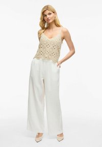 Top de tirantes beige de ganchillo con patrones circulares, combinado con pantalones anchos blancos. La modelo está de pie con una mano en el bolsillo, llevando tacones de color beige.