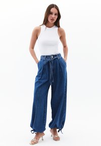 Housut, joissa on leveät lahkeet ja nauha vyötäröllä, tummansiniset. Ominaisuuksiin kuuluu kaksi etupokketa ja säädettävät nauhat lahkeensuussa. Yhdistetty valkoiseen toppiin.