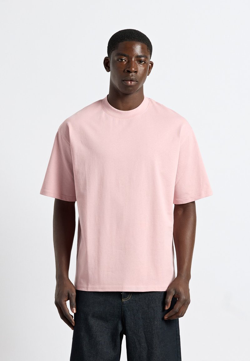 Jeune homme portant un t-shirt ample rose clair et un pantalon foncé, debout devant un fond blanc uni, faisant face.