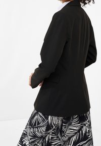 Černý blazer s dlouhými rukávy a přiléhavým střihem, se švem na zadní straně. Nosený přes vzorovanou sukni v černo-bílé barvě.