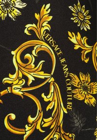 Tessuto nero con motivi floreali gialli e barocchi, con il testo "VERSACE JEANS COUTURE" in un font elegante.