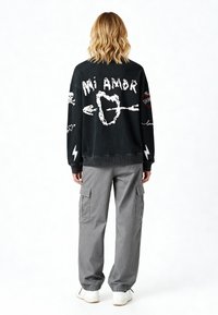 Un sweat-shirt noir avec du texte graphique blanc "Mi Amor" et des motifs, associé à un pantalon cargo gris. Il présente des poignets élastiques et des poches latérales.