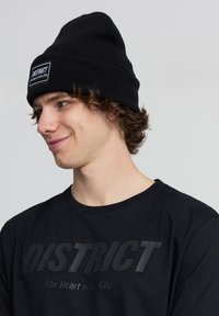 Il berretto di lana nero presenta un'etichetta frontale con "DISTRICT" e "Il cuore della città". Il modello indossa una maglietta nera con un logo prominente.