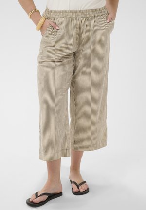 Beige and white vertical striped cropped pants worn with black flip-flops, wrists with chunky bracelets, hands in pockets.