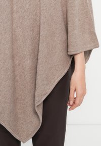 Poncho de malha bege com uma forma solta e assimétrica. Apresenta uma textura suave e mangas largas, combinado com calças castanhas escuras.