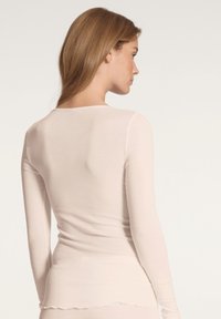 Haut rose clair à manches longues avec un col rond, texture côtelée et ourlet en forme de vagues. Le matériau semble doux et ajusté.