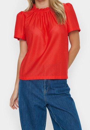 Vrouw die een felrode korte mouwen blouse met een geplooide hals draagt en blauwe spijkerbroek, staand met één hand in de zak.