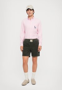 Chemise boutonnée rose clair avec des détails brodés, associée à un short en velours côtelé vert foncé, une casquette grise et des mocassins beige avec des chaussettes blanches.