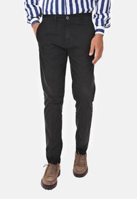 Pantaloni neri in cotone con vestibilità slim, dotati di chiusura con bottone, passanti per cintura e tasche laterali, abbinati a scarpe marroni con lacci.