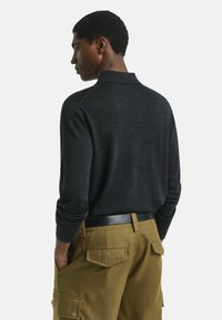 Man in een donkerkleurig shirt met lange mouwen en olijfgroene cargobroek met zakken, staand met handen in zijn zakken, met zijn rug naar de camera tegen een effen achtergrond.