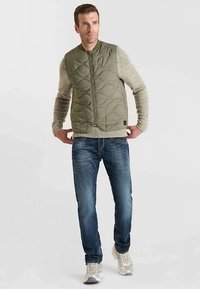 Gewatteerd olijfgroen vest met afgeronde vormen, een ritssluiting aan de voorkant en mouwloos, gedragen over een lichtgrijze trui en blauwe spijkerbroek, afgewerkt met grijze sneakers.