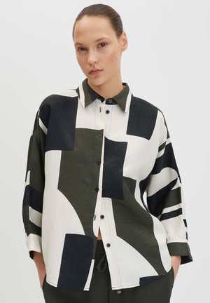 Femme aux cheveux attachés portant une chemise boutonnée à manches longues avec de grands motifs géométriques noirs, blancs et olive sur un fond uni.