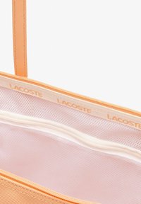 Bolsa de mano naranja con material liso y texturizado. El interior presenta un forro de malla clara y un bolsillo con cremallera. Marca Lacoste exhibida.