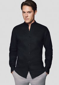 Czarna, długa koszula z mandaryńskim kołnierzykiem, wykonana z teksturowanej tkaniny i zapinana na guziki z przodu. Noszona z jasnymi spodniami eleganckimi.