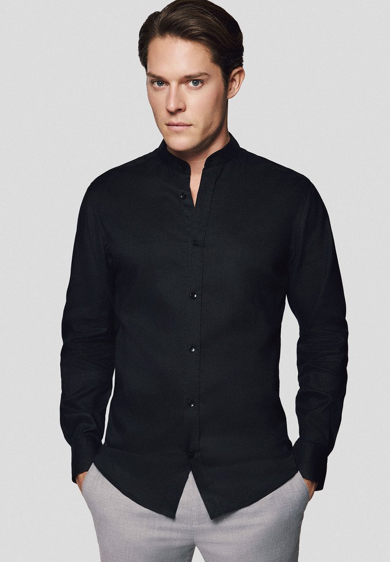 Czarna, długa koszula z mandaryńskim kołnierzykiem, wykonana z teksturowanej tkaniny i zapinana na guziki z przodu. Noszona z jasnymi spodniami eleganckimi.