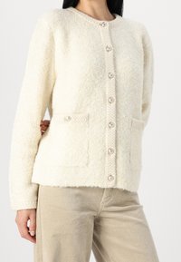 Krämmfärgad cardigan i en texturerad, lurvig tyg, med rund halsringning, två framfickor och dekorativa knappar.