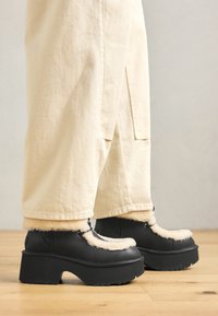 Sorte plattformstøvler med rund tå, som har en fluffy kremfarget krage og snøre detaljer, kombinert med løse beige bukser på et tregulv.