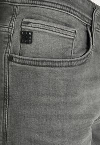 Detailní záběr zadní kapsy šedých džín z denimu s malým černým textilním štítkem s bílými písmeny a prošitými švy.