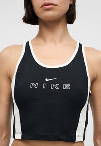 Γυναίκα που φοράει μαύρο και λευκό crop top από τη Nike με το λογότυπο και κείμενο της Nike στο κέντρο του στήθους, μπροστά σε απλό φόντο.