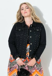 Svart denimjacka med silverfärgade nitar på framsidan, med krage och knäppning, tillsammans med en mönstrad klänning i orange och svart.