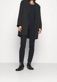 Person som bär en svart oversized blazer, mörkblå rutig slim-fit byxor, svarta ankelskor och ett kraftigt guldhalsband.