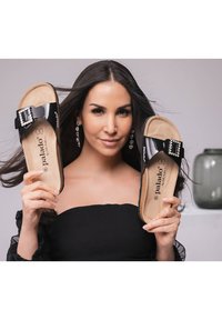 Zwarte gepatenteerde slidesandalen met een grote rhinestone gespsluiting, een beige gevoerde voetbed en "Palado" branding op de inlegzool, vastgehouden door een persoon.