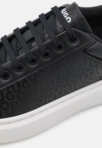 Baskets noires avec une tige texturée présentant un grand motif de logo en relief, des lacets noirs et une semelle en caoutchouc blanche. Éléments de design minimalistes.