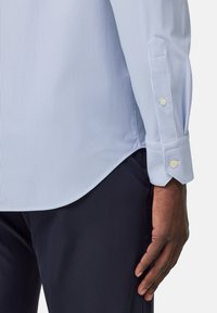 Camicia a righe blu chiaro con maniche lunghe, polsini abbottonati e orlo curvo. Il tessuto appare liscio con una trama fine. Pantaloni scuri visibili.