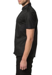 Camisa negra de manga corta con cuello puntiagudo, parte delantera con botones, bolsillo en el pecho izquierdo y una textura suave. Llevada por un modelo masculino de perfil.