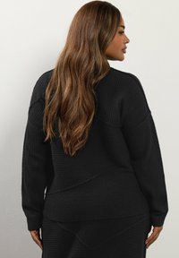 Svart stickad tröja med ribbad textur, rund halsringning och långa ärmar. Geometriska sömmar ger visuell intresse till designen.