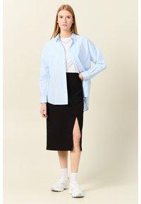 Ljusblå randig oversize-skjorta, vit t-shirt, svart kjol med sidoklyvning och vita sneakers, mot en neutral bakgrund.
