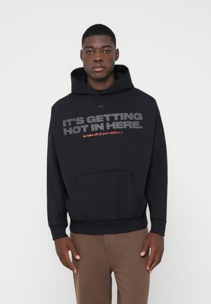 Mlad moški nosi črno kapuco z napisa "POSTAJA PARENO V TEM PROSTORU. zato slecite vse obleke ;)", in rjave hlače, stoji ob belem zidu.