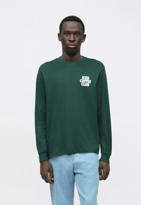 Tamnozeleni dukserica dugih rukava sa bijelim natpisom "ICED COFFEE CLUB" na lijevoj strani prsnog dijela, izrađena od mekog materijala s klasičnim krojem.