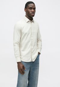 Svetlo béžová košeľa s dlhými rukávmi, golierom na gombíky, textúrovanou látkou a jemným logom. Kombinovaná s voľnými modrými džínsami.