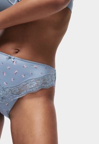 Intimo in pizzo blu chiaro con motivi floreali rosa. Presenta una delicata rifinitura in pizzo e un piccolo dettaglio a fiocco nella parte anteriore. Texture liscia.