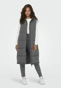 Gilets rembourrés longs et gris avec capuche, dotés d'une fermeture éclair, de poches latérales et d'un design matelassé. Porté par-dessus un haut blanc et un pantalon gris.