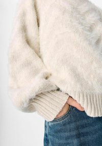 Krēmkrāsas džemperis ar pūkainu struktūru, kas raksturojas ar riboto aproci un brīvu piegriezumu, valkāts kopā ar zilām džinsa biksēm.